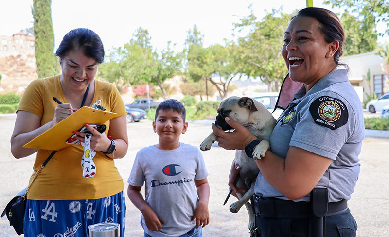 A smiling woman fills out information on a clipboard as Christina Avila holds a small dog and a boy looks on.