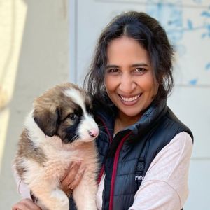 Jyothi smiling, wearing a blue parka, holding a fluffy puppy