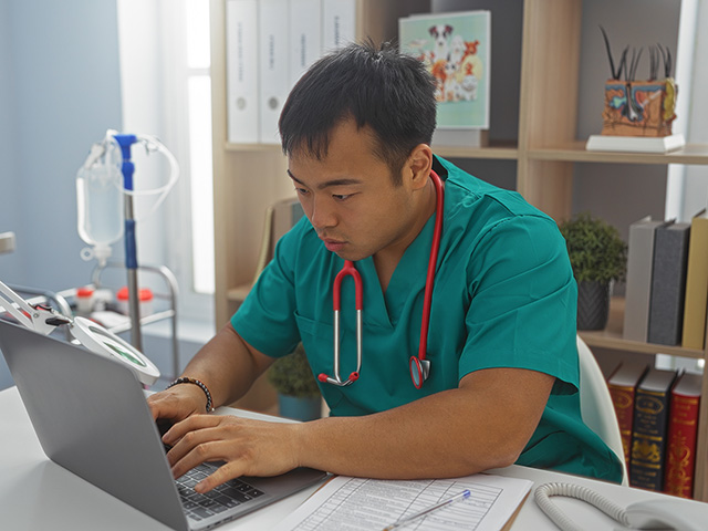 Asian veterinarian wearing turquoise scrubs sitting at desk in office with stethoscope hanging around his neck types on a laptop computer