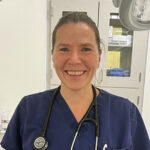 Close-up of a smiling Doctor Beck wearing dark blue scrubs, with a stethescope hanging around her neck