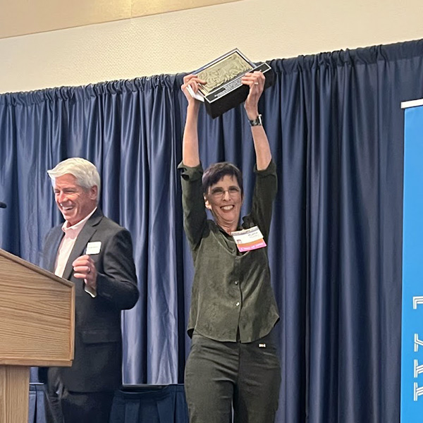 Mark D. Stetter, Dean, at podium on the left, smiling broadly. Dr. Hurley smiles and holds aloft the plaque signifying the award.